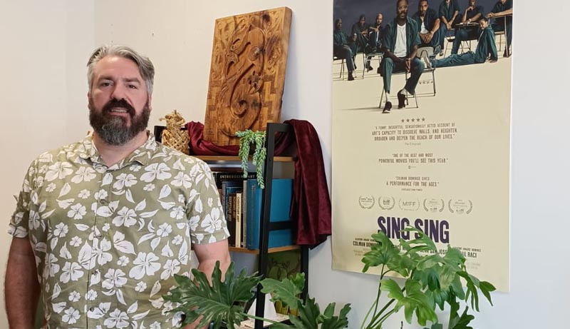Neil Wallace in his office, standing in front of a small caribg and a poster on the wall advertising Sing Sing, the movie